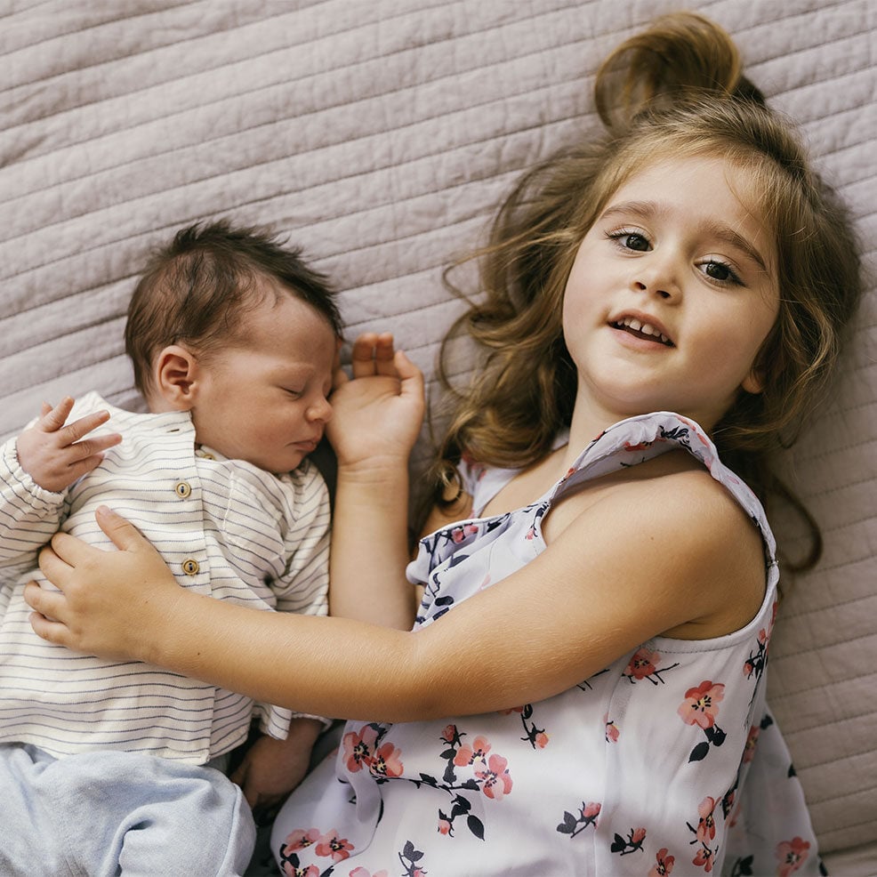 Ein kleines Mädchen liegt mit einem schlafenden Baby auf einer Decke und lächelt. 